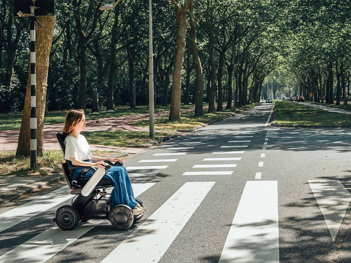 Frau in WHILL Model C2 Elektrorollstuhl überquert einen Zebrastreifen auf einer baumgesäumten Straße.