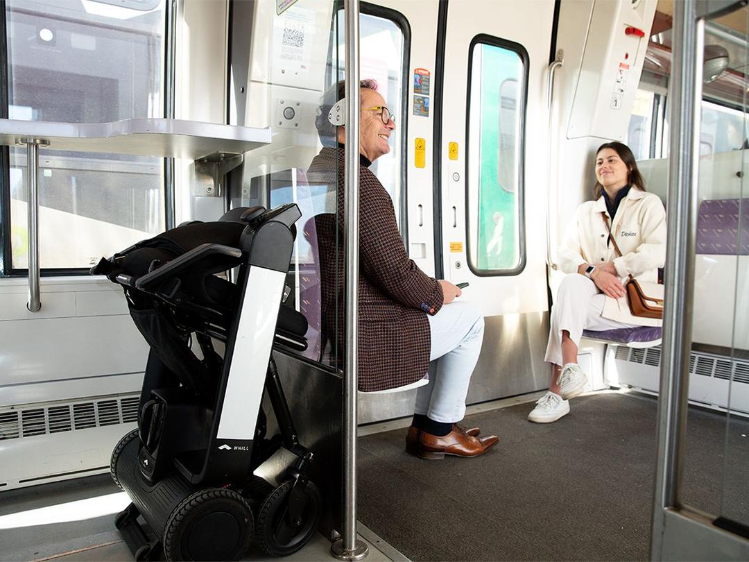 Ein Mann und eine Frau sitzen in einem Zugabteil. Hinter dem Mann ist hinter einer Glaswand ein zusammengeklapptes WHILL Model F verstaut.