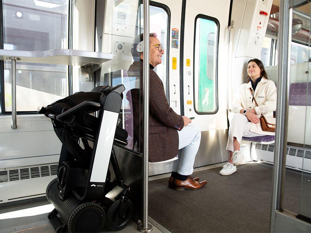 Ein Mann und eine Frau sitzen in einem Zugabteil. Hinter dem Mann ist hinter einer Glaswand ein zusammengeklapptes WHILL Model F verstaut.