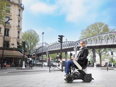 Frau im Elektrorollstuhl WHILL Model C2 steht auf einem Gehweg, im Hintergrund eine Ampel in der Stadt.