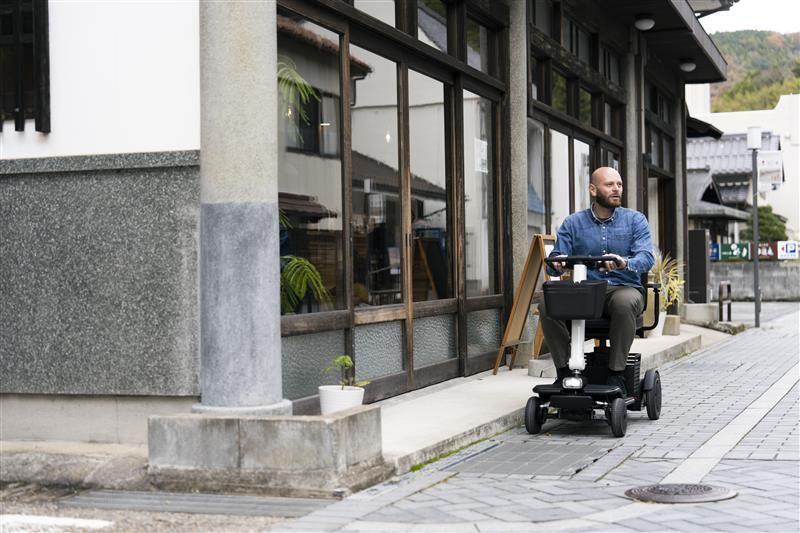 Ein Mann auf einem weißen Scooter fährt an einer Hauswand mit Fenstern entlang.