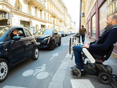 Mann im Elektrorollstuhl WHILL Model C 2 steht auf einem Gehweg an der Straße im städtischen Straßenverkehr.