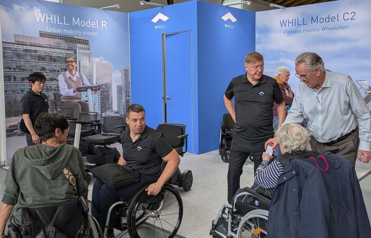 WHILL-Messestand in blau auf einer Messe, mehrere WHILL-Mitarbeiter betreuen Messebesucher in Rollstühlen.