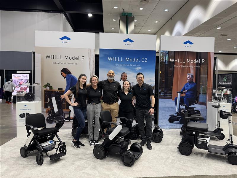 A smiling group of five people stands in front of WHILL power chair and mobility scooter display at an indoor exhibition. Three mobility models are showcased.