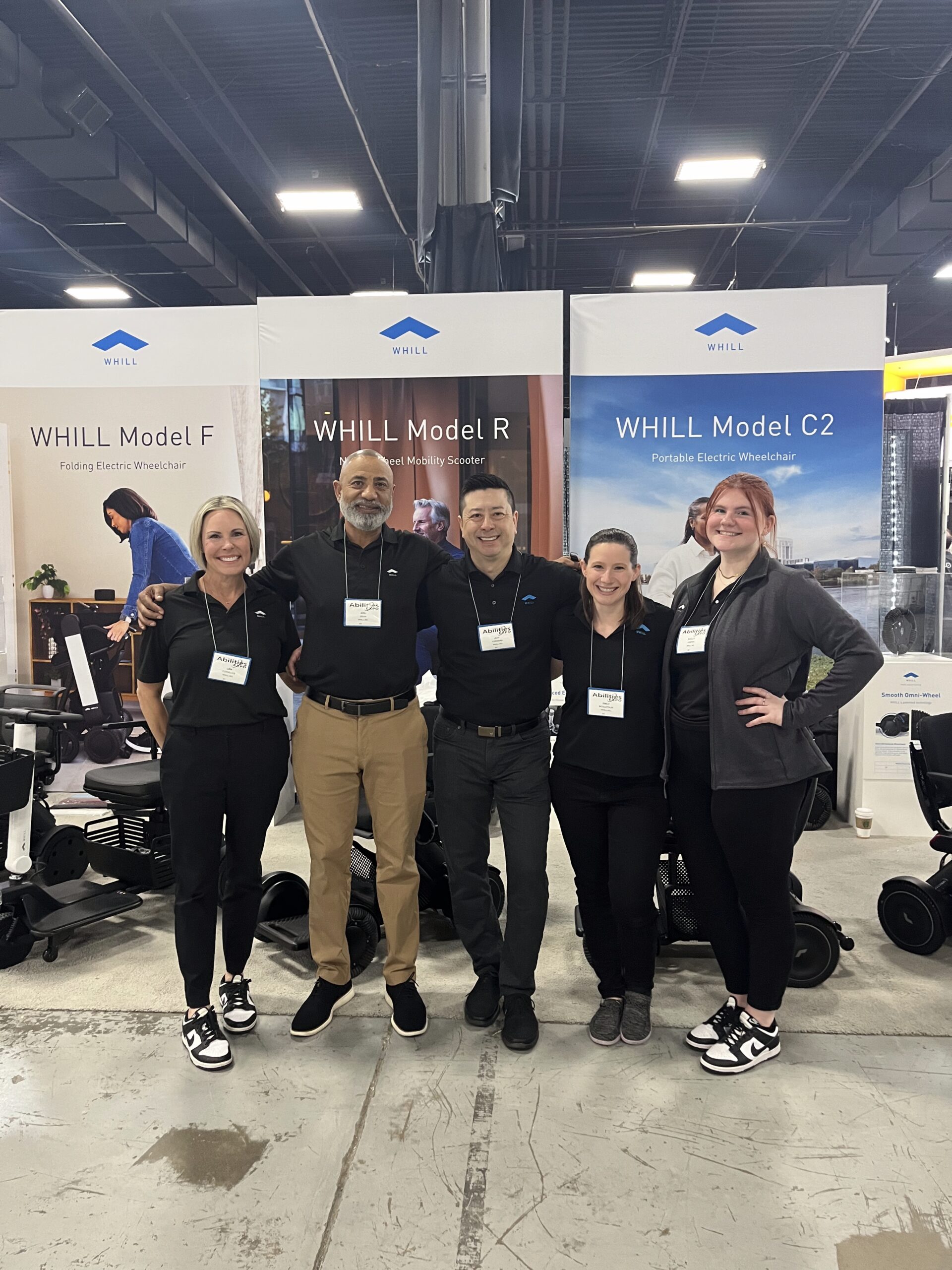 Five people wearing black shirts, smiling and standing in front of banners for WHILL mobility devices. The atmosphere is friendly and professional.