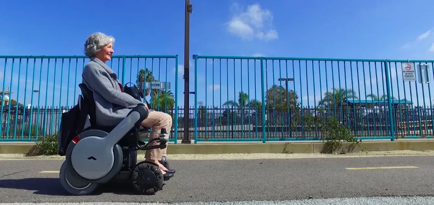 An elderly woman smiles while riding a WHILL Model A Power Chair on a sunny day. A blue fence and palm trees in the background suggest a relaxed atmosphere.