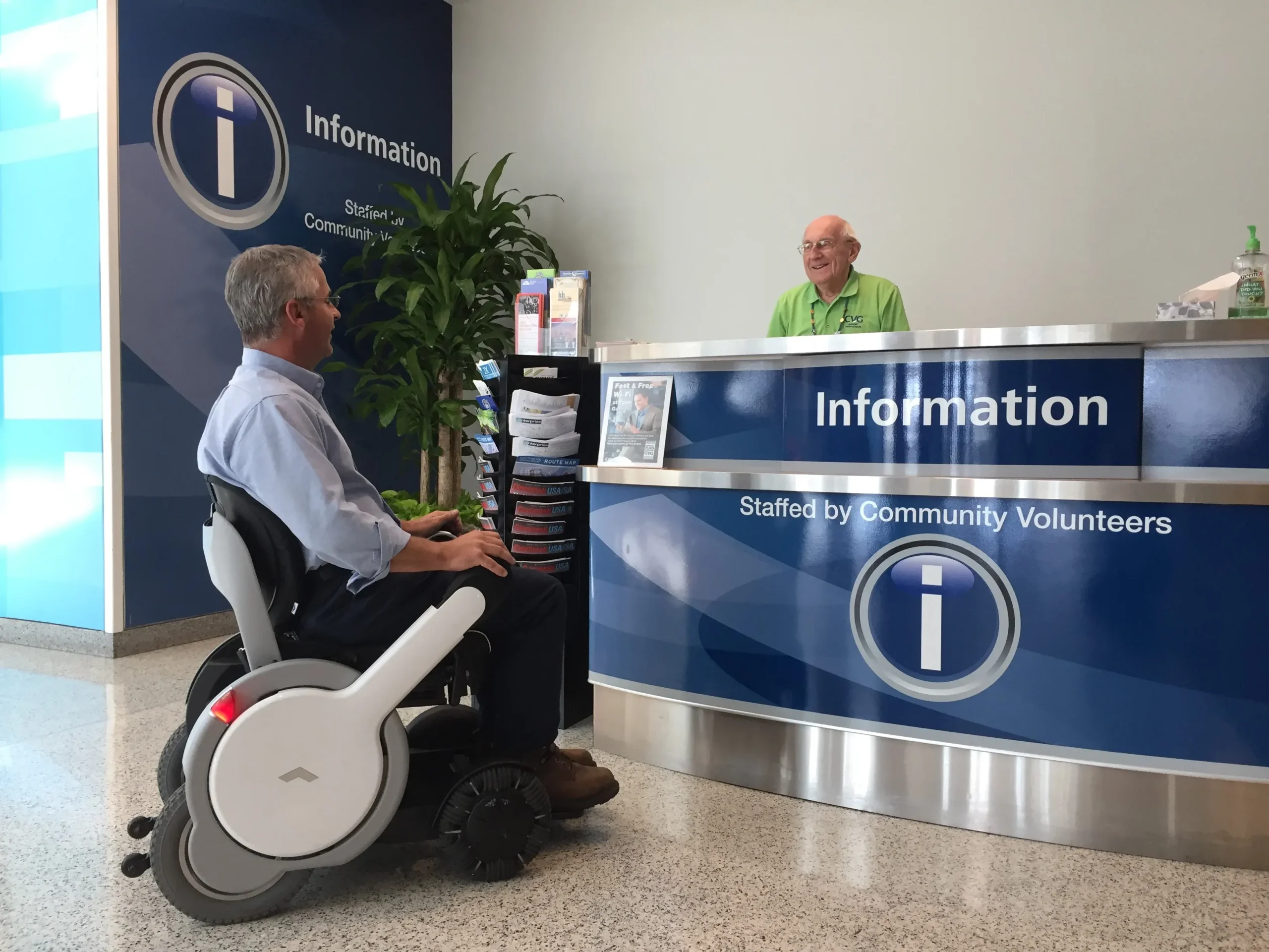 A man in a WHILL Power Chair speaks with a smiling volunteer at an information desk. The desk is blue, with signs reading "Information" and "Staffed by Community Volunteers."