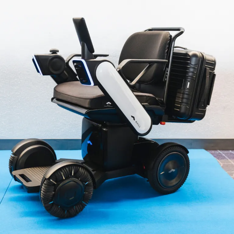 A WHILL autonomous service power chair with a suitcase attached at the back. It sits on blue floor.