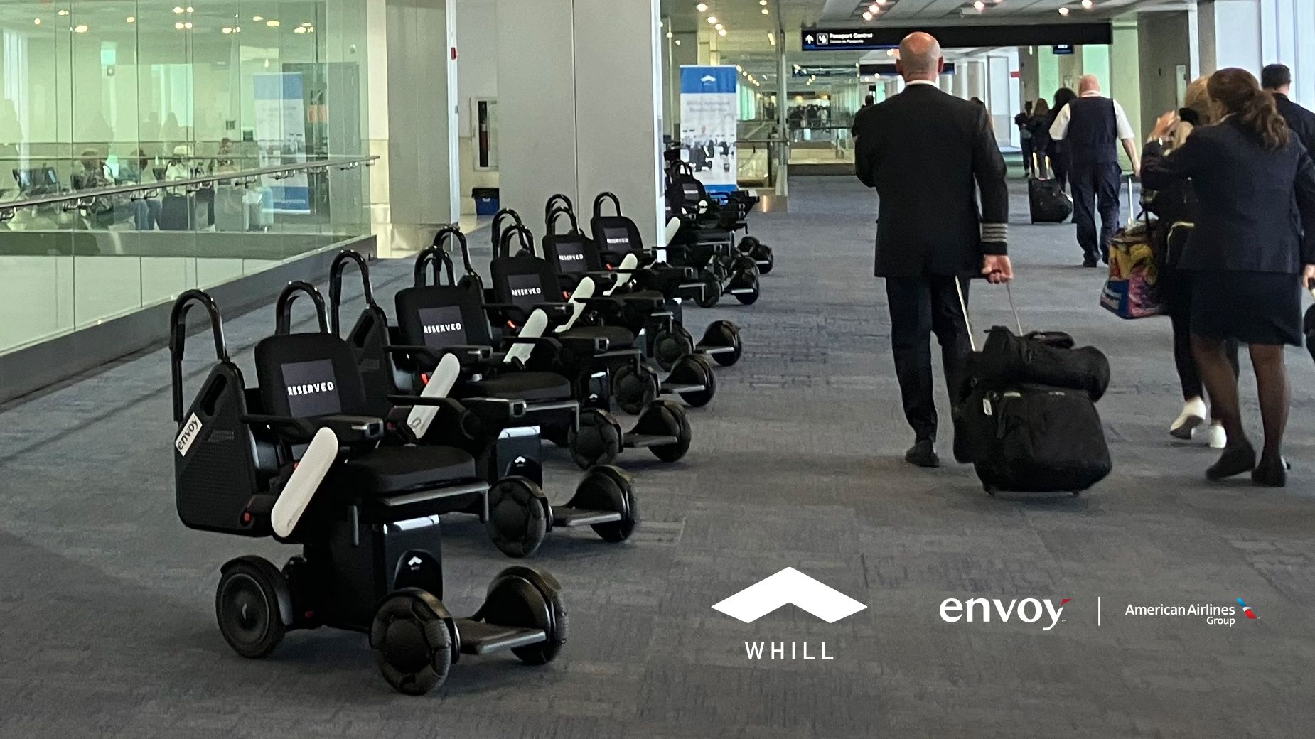 Row of sixWHILL Autonomous Service Power Chairs lined up in an airport corridor. People walk by with luggage, creating a dynamic airport ambiance. Logos for WHILL, Envoy, and American Airlines are visible.