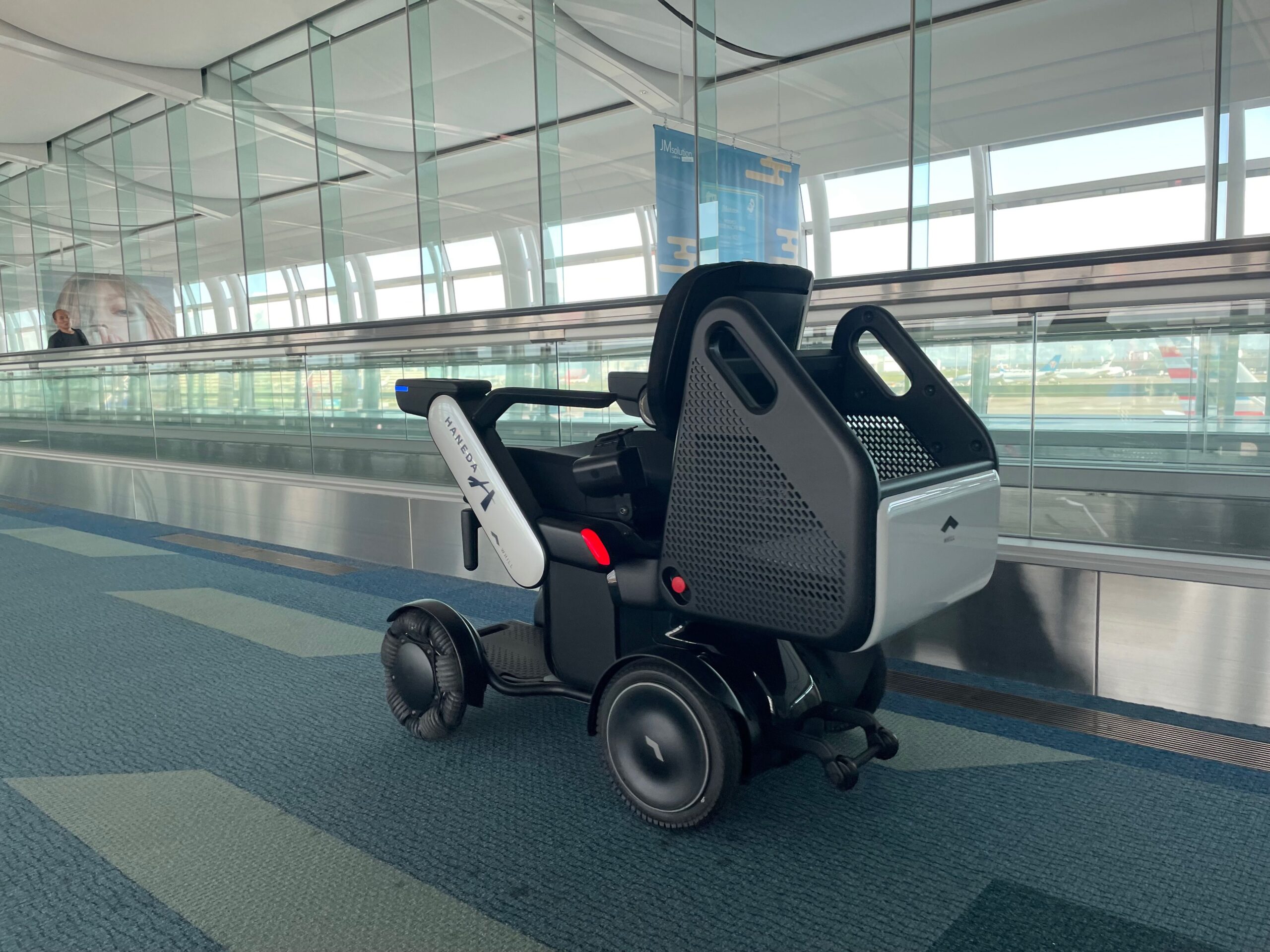 A WHILL autonomous service power chair is positioned in an airport corridor next to large windows, conveying accessibility.
