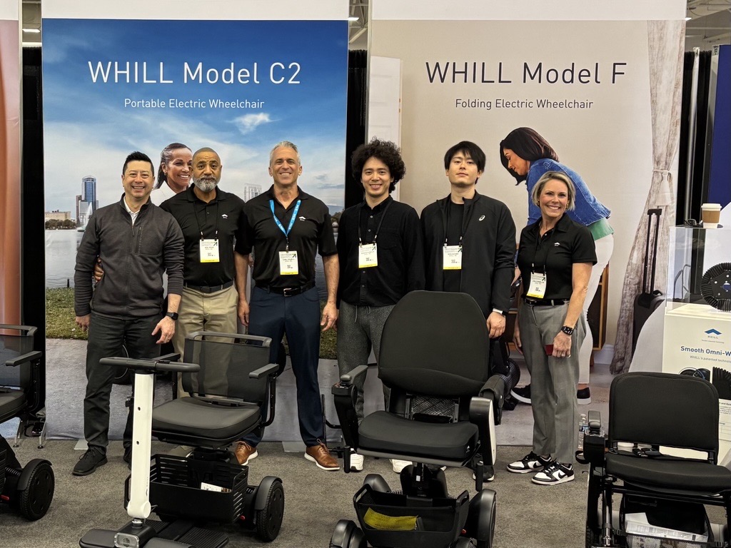 A group of six people stand smiling in front of banners for WHILL Model C2 and Model F electric wheelchairs at a trade show, conveying a professional and friendly atmosphere.