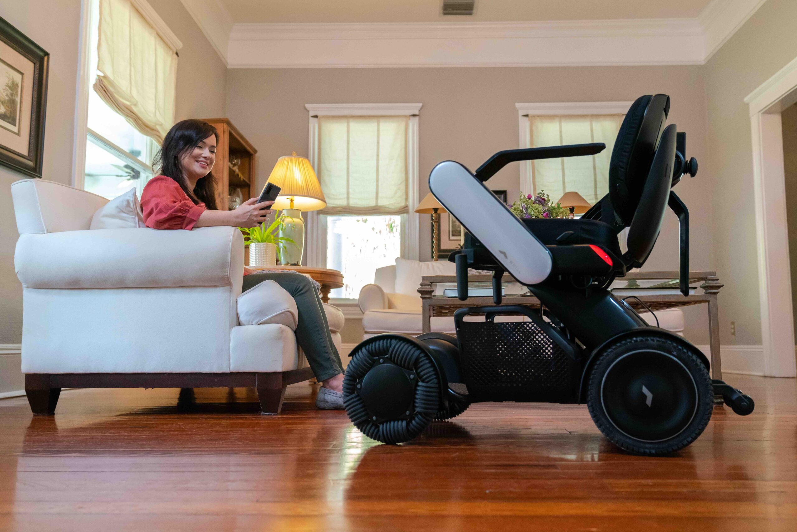 A woman sits on a white sofa in a cozy living room, smiling at her phone. Nearby, a WHILL Model C2 power chair is parked on the wooden floor.