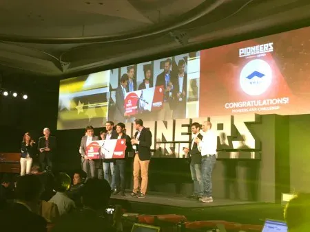 A group on stage celebrates a win at a tech event. They hold a large red award sign, while a screen behind displays "Pioneers" and "Congratulations!"