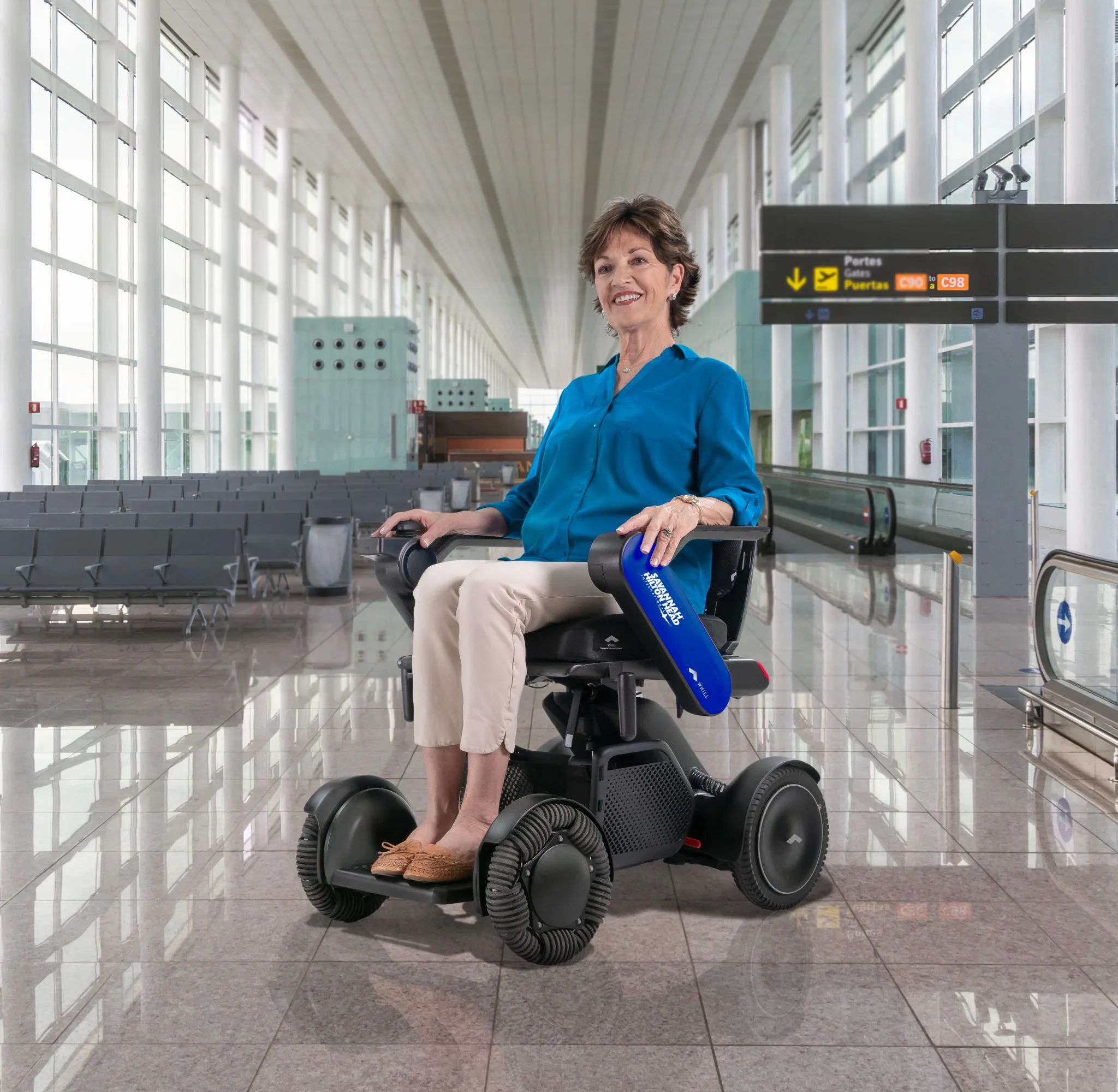 Elderly woman in a blue blouse smiles while seated in a WHILL Autonomous service power chair at a spacious, empty airport terminal. Direction signs visible overhead.