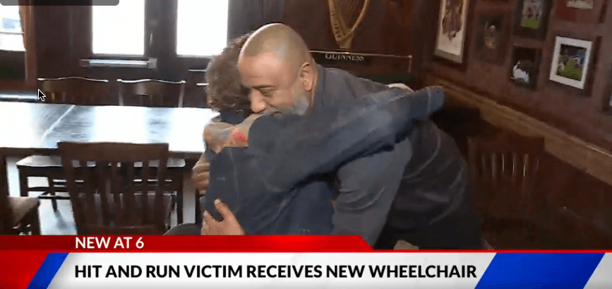 Two people warmly embrace in a cozy room with wooden decor. The atmosphere is joyful. A news banner below reads, "Hit and Run Victim Receives New Wheelchair."