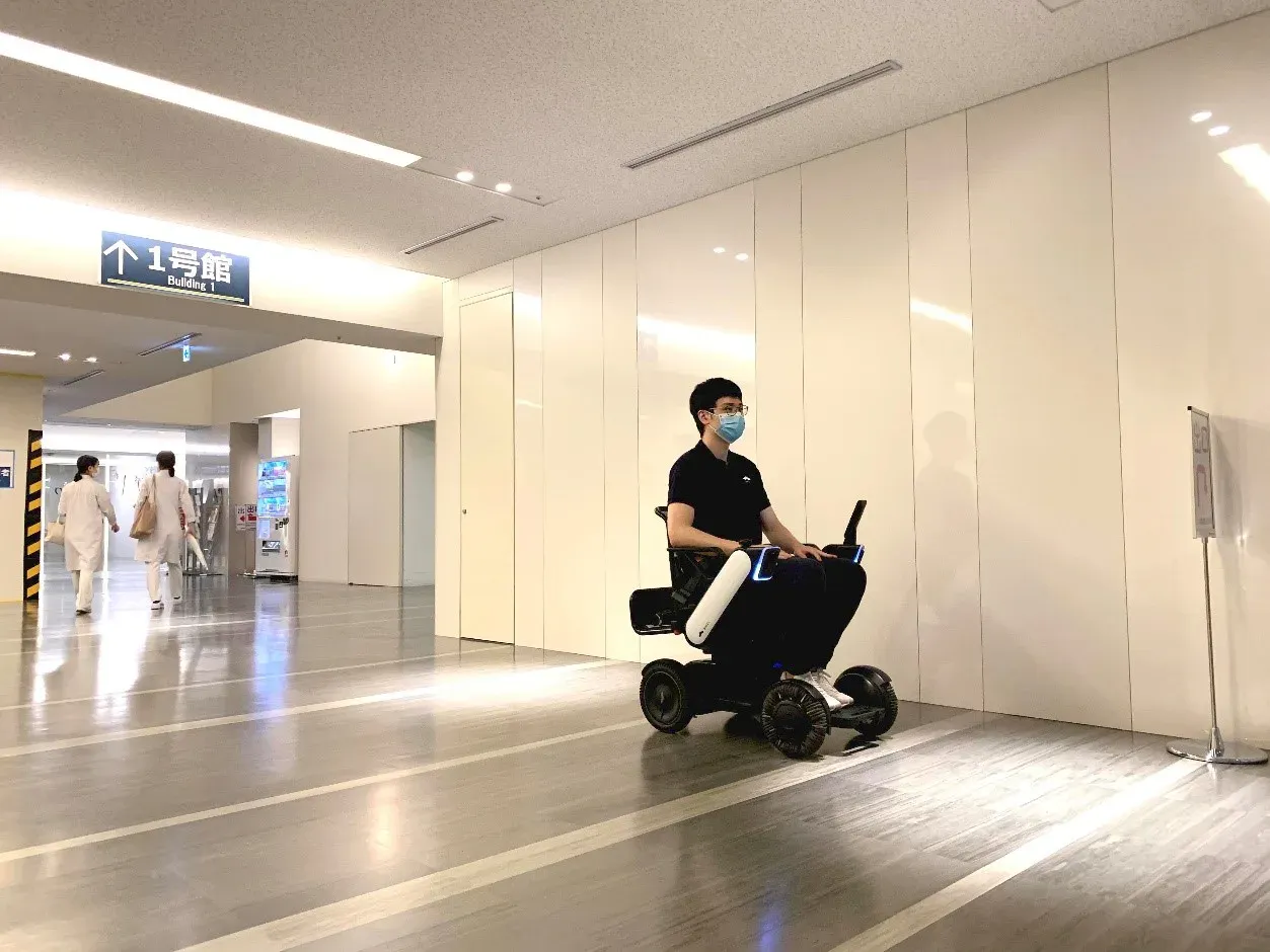 A man wearing a mask rides an WHILL Autonomous service power chair down a bright, modern hallway, creating an atmosphere of accessibility and movement.