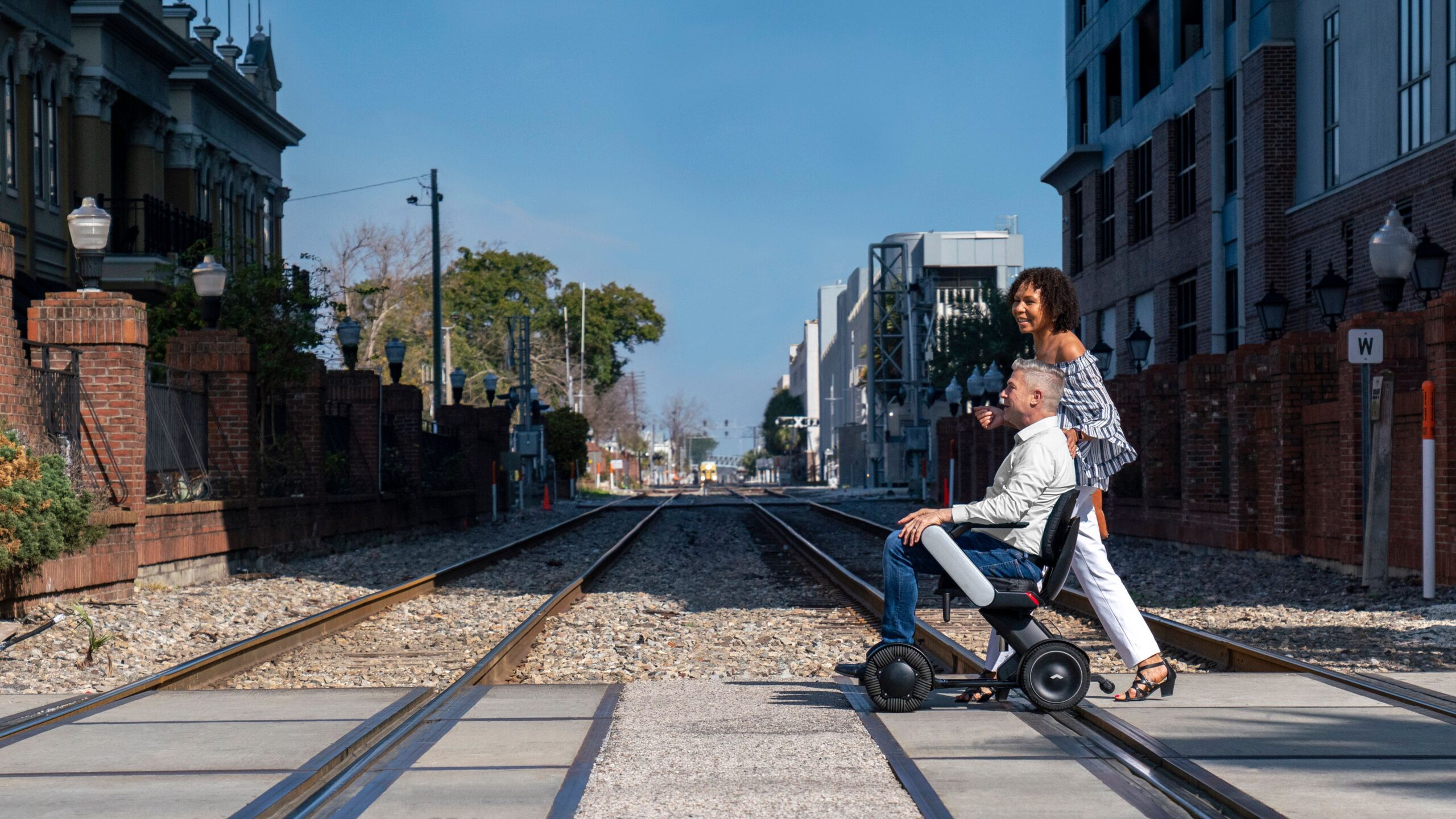A man in a white shirt rides an WHILL Model C2 mobility chair labeled "BIRD" alongside a woman smiling, wearing an off-shoulder top and white pants. They are near a staircase.