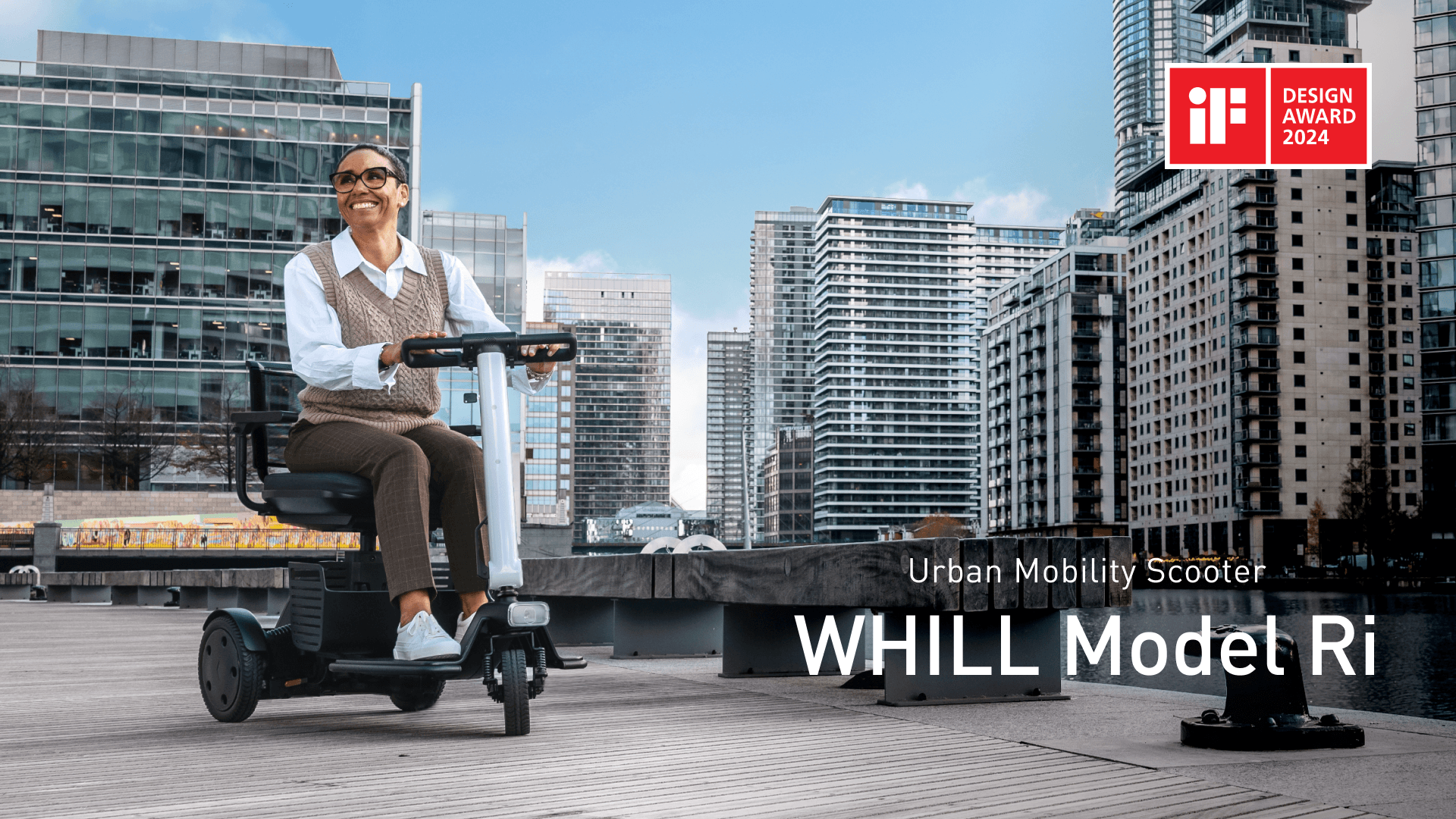 A person joyfully rides a WHILL Model Ri urban mobility scooter on a boardwalk. Modern cityscape in the background. "IF Design Award 2024" logo is visible.