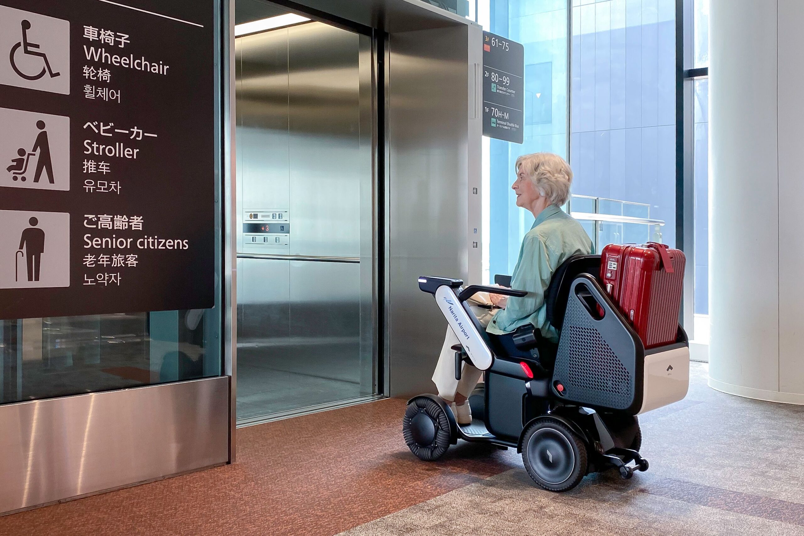 An elderly person in a WHILL Autonomous service power chair waits at an elevator with a suitcase attached. Signs indicate wheelchair access and facilities for seniors.