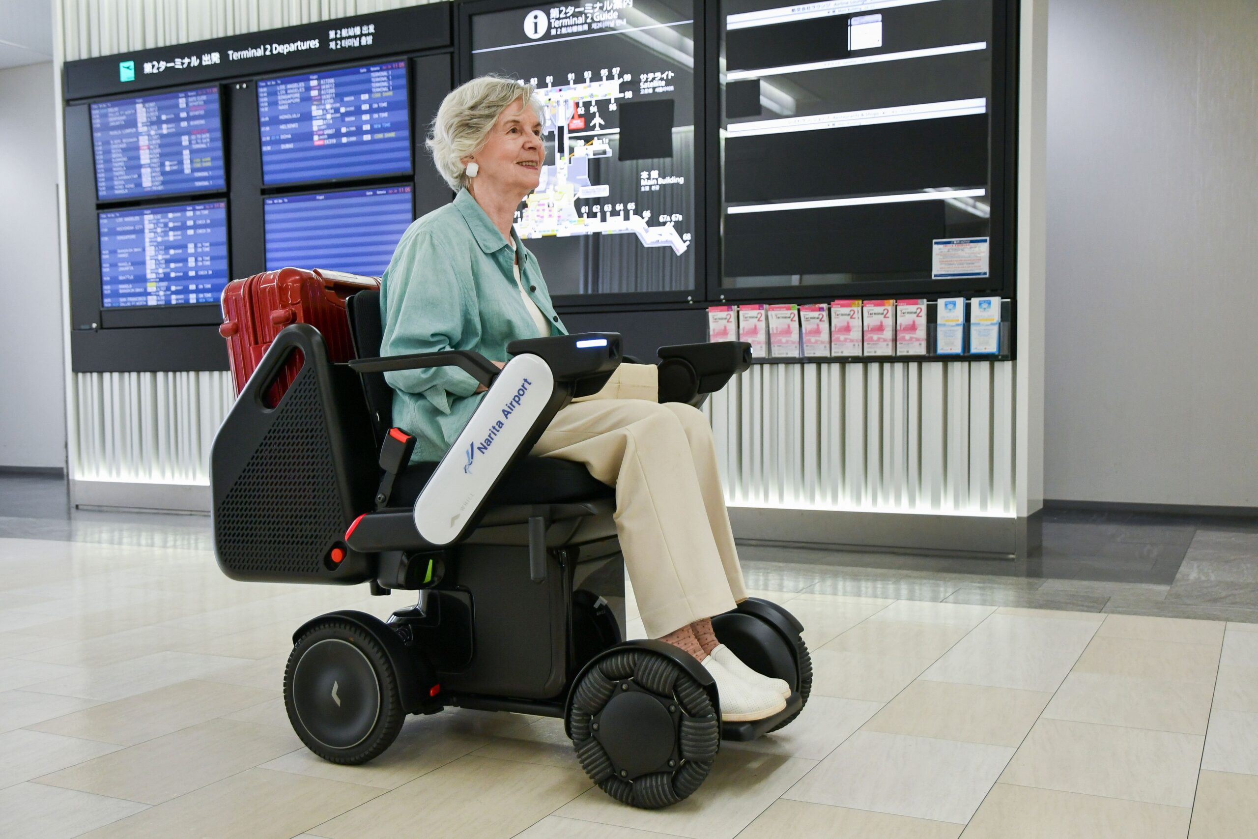An elderly woman smiles while riding an WHILL autonomous service power chair at an airport. She carries a red suitcase. Flight information screens are visible behind her.
