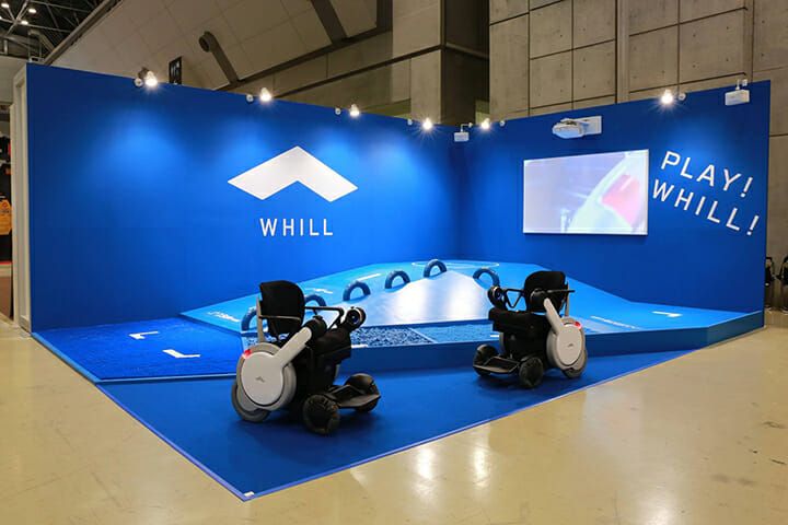 Exhibit booth with two modern Power Chairs from "Whill" brand on a blue platform. The walls display the logo and slogan "Play! Whill!" with spotlights.