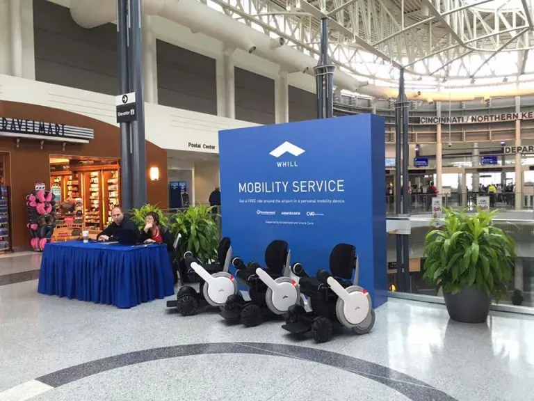 Airport terminal with a "Mobility Service" display featuring personal WHILL Power Chair mobility devices. A table with staff is nearby.