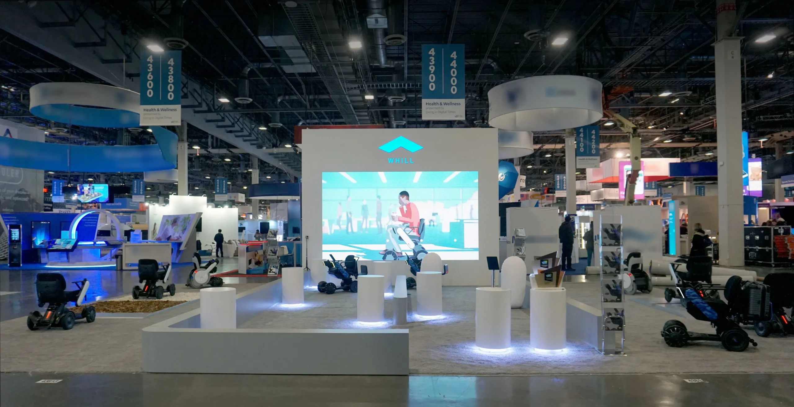 A exhibition booth features a large screen displaying an animated video. Power chairs are positioned around white pedestals under blue lighting.