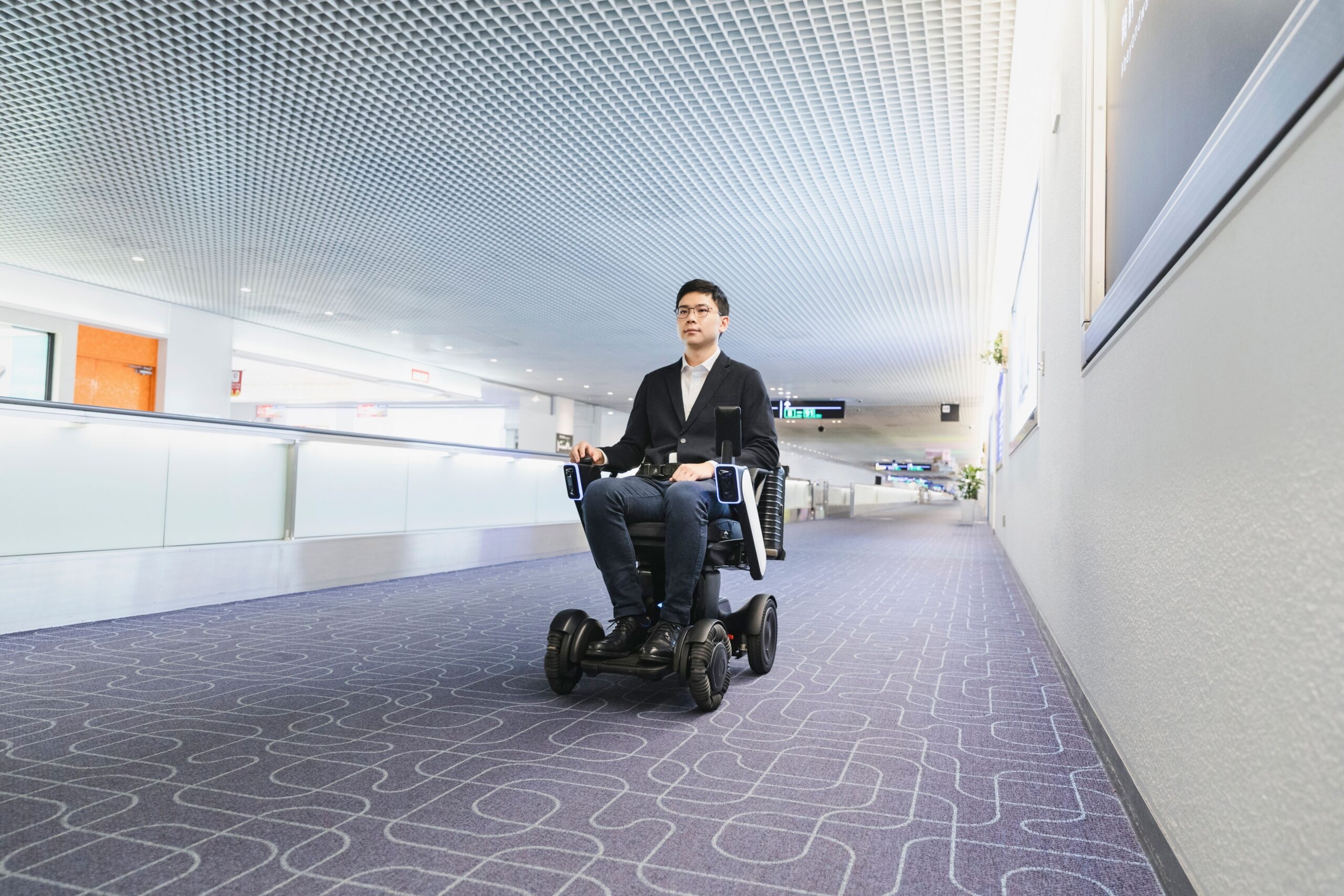 A person in a suit uses an WHILL autonomouse service mobility device in a spacious, modern hallway with patterned carpet.
