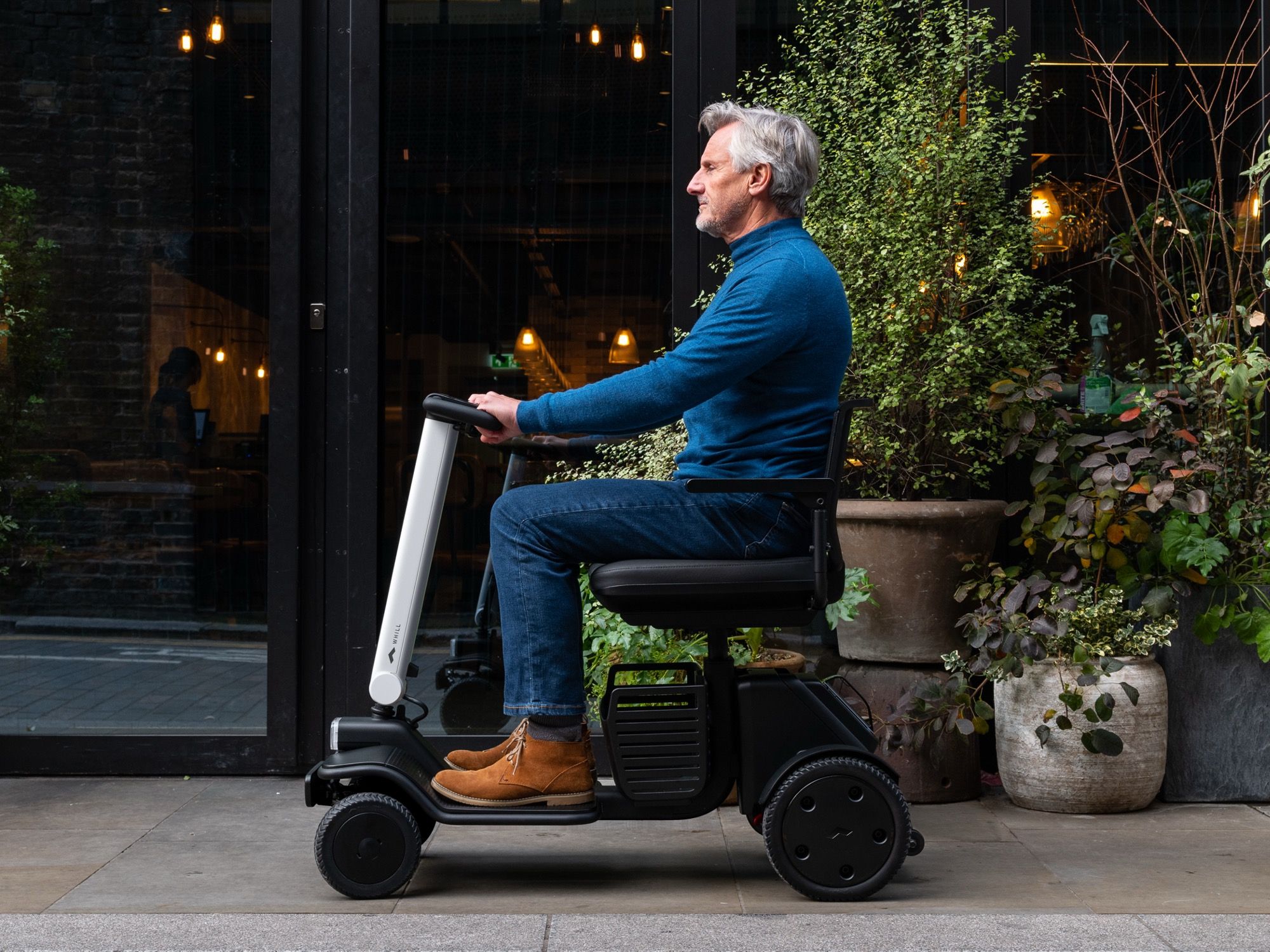 A man rides a mobility scooter on a sidewalk by a modern building. He's wearing a blue sweater and jeans. Green plants are in pots nearby, conveying calm.