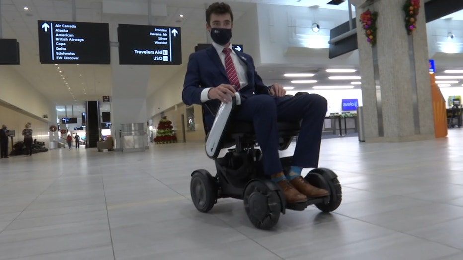 A man in a blue suit and red tie, wearing a face mask, rides a WHILL autonomous service power chair in an airport. Signs for airlines hang above.