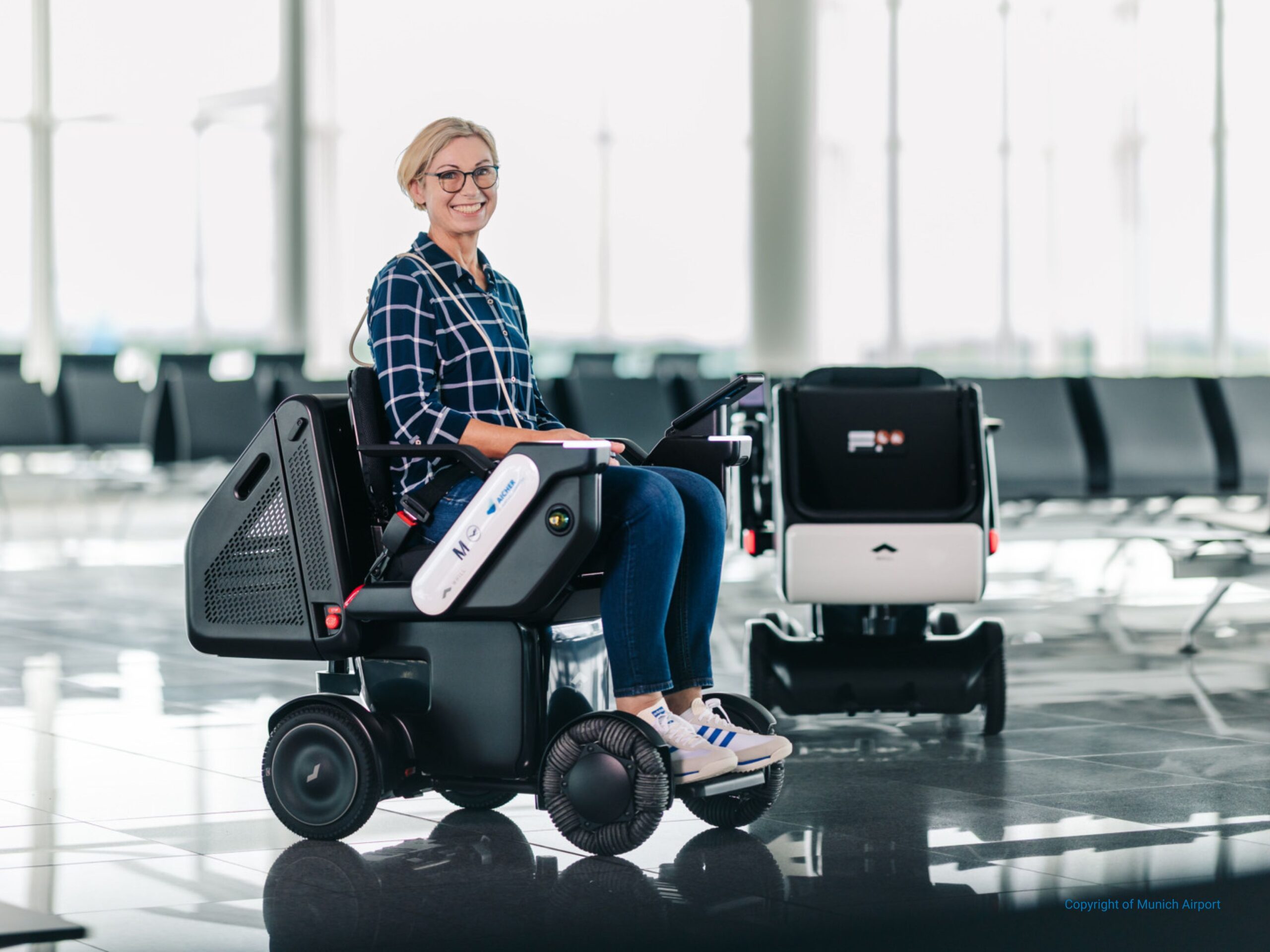 A smiling person sits in an airport on a WHILL autonomous service power chair, wearing casual clothes. The setting is bright and spacious, conveying a positive mood.
