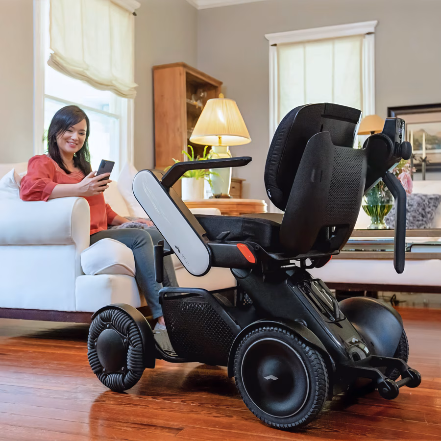 A woman is sitting on a couch with a phone next to a WHILL power chair.