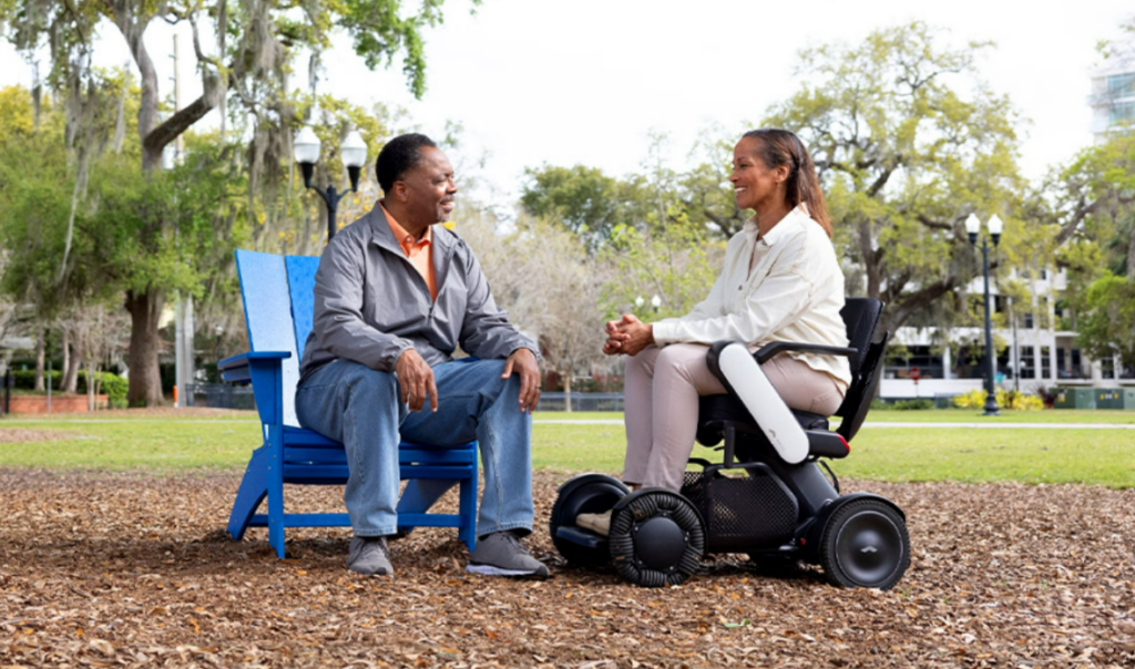 Two people are engaged in a friendly conversation in a park. One sits on a WHILL Model C2 power chair, and the other sits in a blue chair. Trees and a house are in the background.