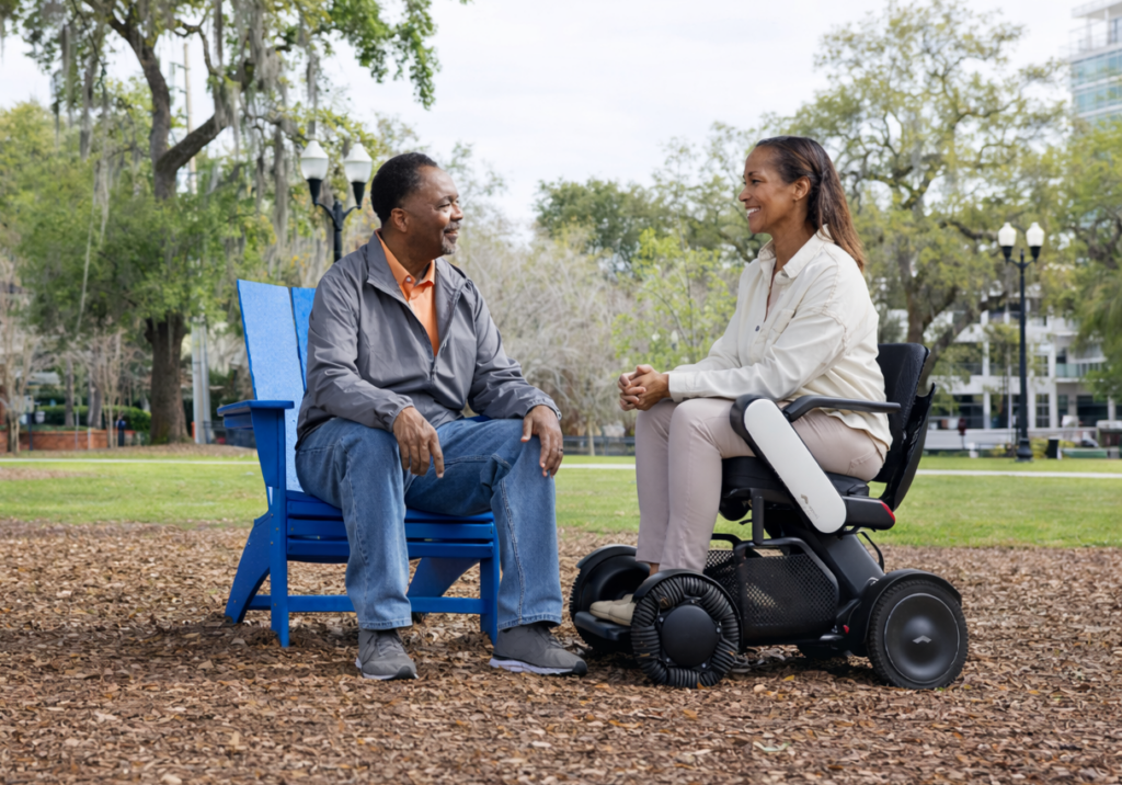 Two people are engaged in a friendly conversation in a park. One sits on a WHILL Model C2 power chair, and the other sits in a blue chair. Trees and a house are in the background.